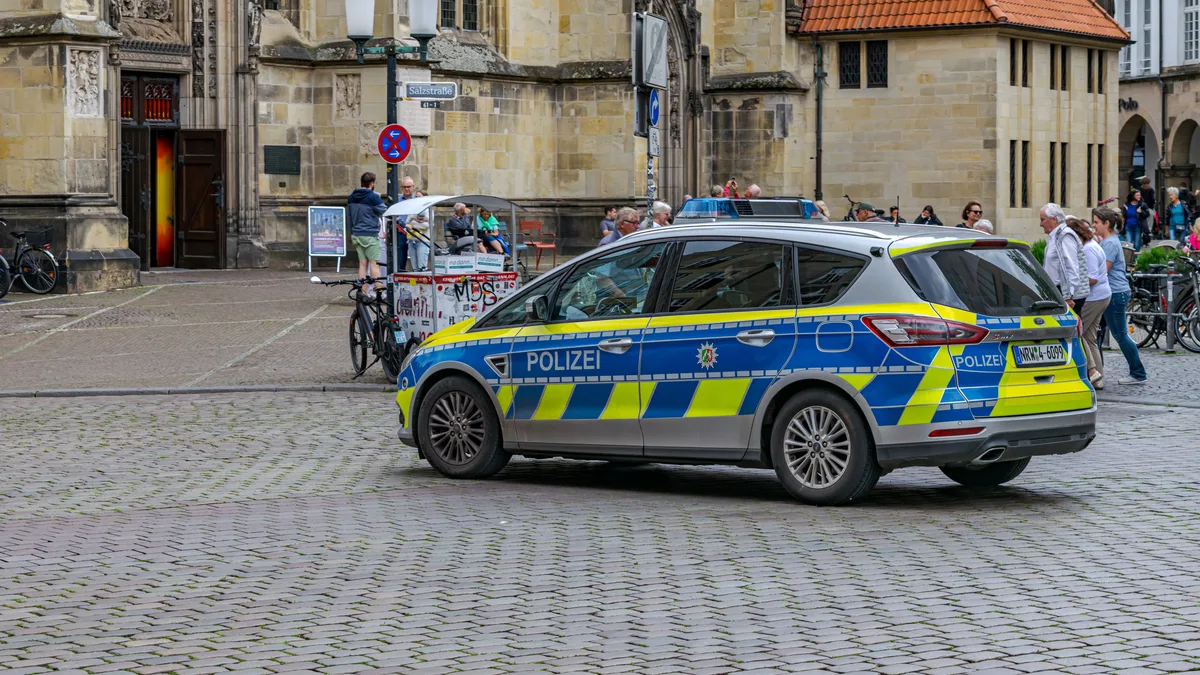 Ein Polizeiwagen steht auf einem historischen Platz in Münster, Deutschland, umgeben von Fußgängern und Fahrrädern.