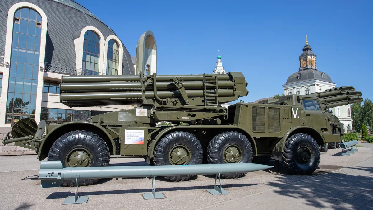 Ein militärischer Raketenwerfer im Freien ausgestellt, mit einer Kulisse aus einzigartiger Architektur und klarem blauen Himmel.