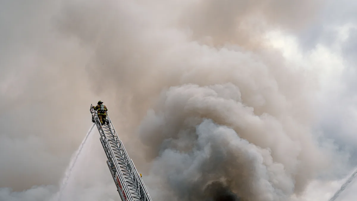 Ein mutiger Feuerwehrmann bekämpft ein riesiges Feuer von einer hohen Leiter aus, umgeben von dichten Rauch.