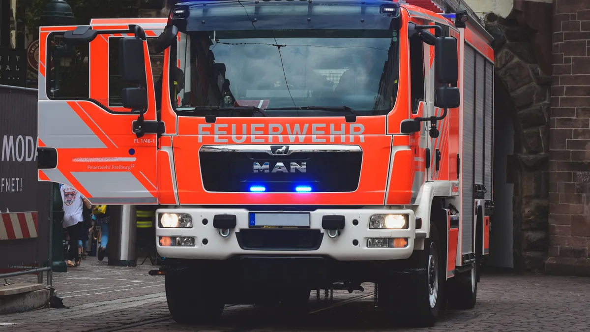 Helles rotes Feuerwehrfahrzeug mit blauen Lichtern auf einer Stadtstraße bei Tageslicht.
