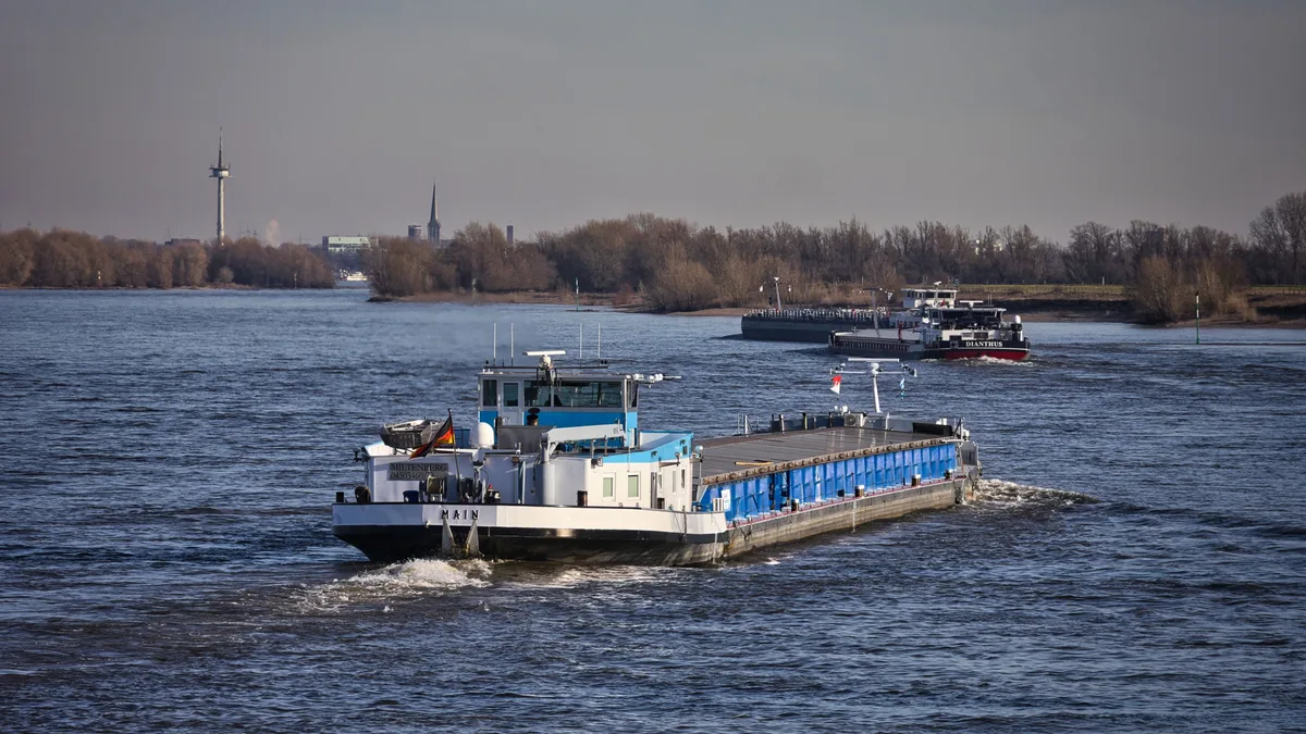 Ein Frachtschiff fährt an einem klaren Tag auf dem Rhein, im Hintergrund ist eine Stadtkulisse zu sehen.