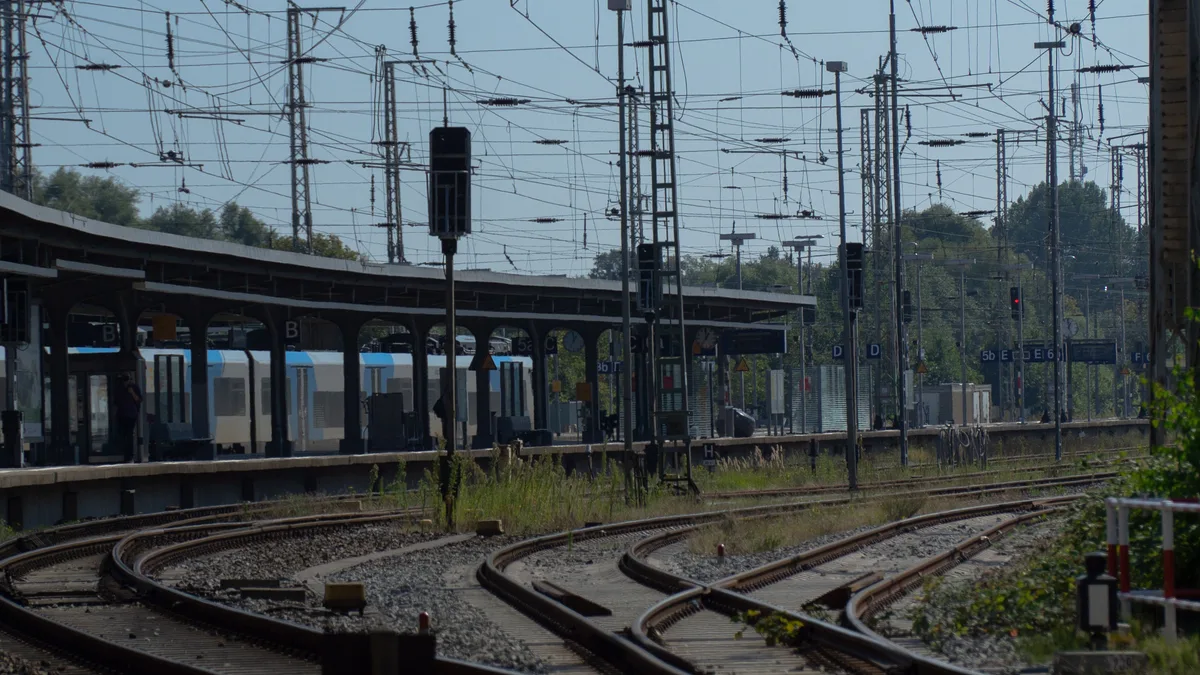 Blick auf mehrere Eisenbahnstrecken, die an einem Bahnhof unter Oberleitungen konvergieren.