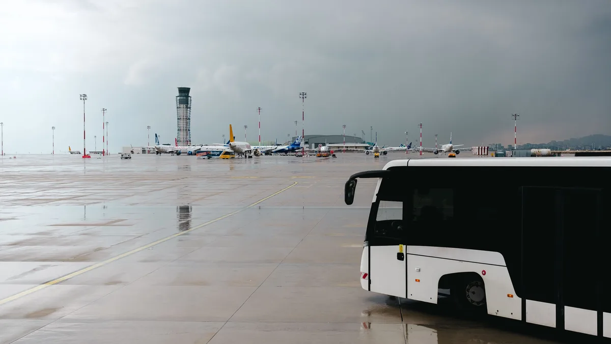 Verregnetes Flughafenrollfeld mit einem Passagierbus und Flugzeugen im Hintergrund.