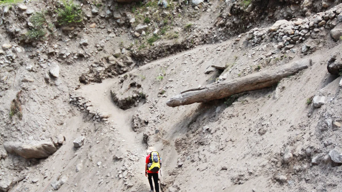 Ein einsamer Wanderer mit gelbem Rucksack durchquert einen unebenen Dirt Path inmitten felsigem Bergterrain.