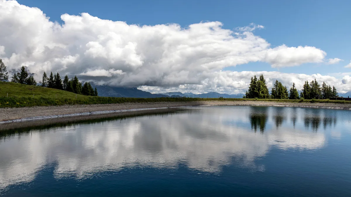 Ein ruhiger See auf dem Land, der dramatische Wolken und Bäume unter einem hellen Himmel spiegelt.