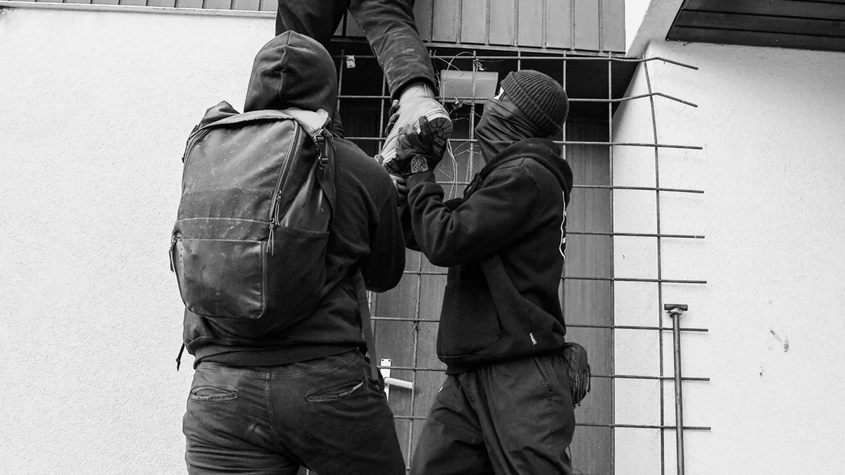 Drei Personen mit Masken, die einen Zaun erklimmen, was in Schwarz-Weiß auf Eindringen hinweist.