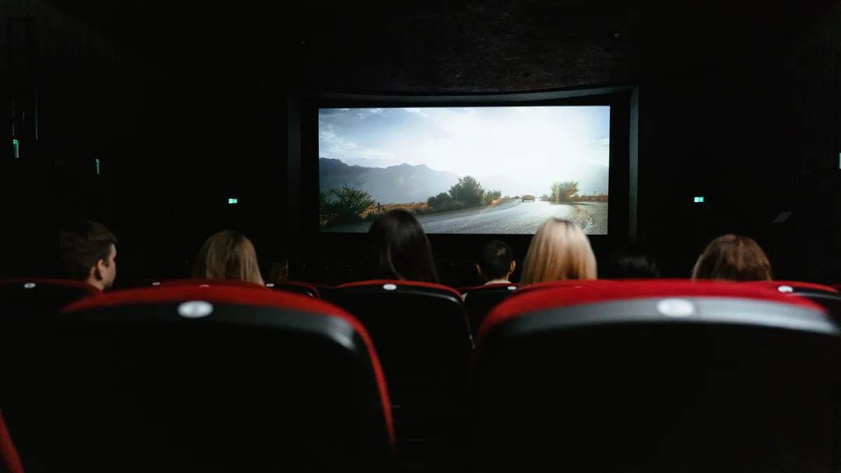 Blick von hinten auf Menschen, die einen Film in einem Kino mit roten Sitzen und einer großen Leinwand ansehen.