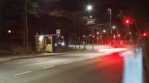 Eine ruhige städtische Straße in der Nacht mit einer Bushaltestelle, Straßenlaternen und Lichtstreifen von vorbeifahrenden Autos.