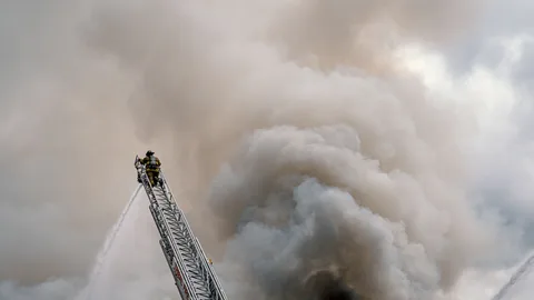 Ein mutiger Feuerwehrmann bekämpft ein riesiges Feuer von einer hohen Leiter aus, umgeben von dichten Rauch.