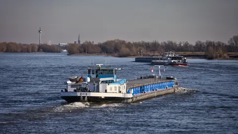 Ein Frachtschiff fährt an einem klaren Tag auf dem Rhein, im Hintergrund ist eine Stadtkulisse zu sehen.