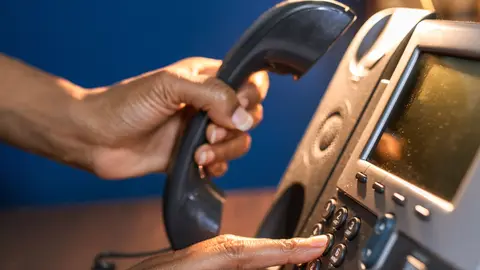 Eine Person wählt eine Telefonnummer an einem Tischtelefon, mit dem Fokus auf die Handdetails.