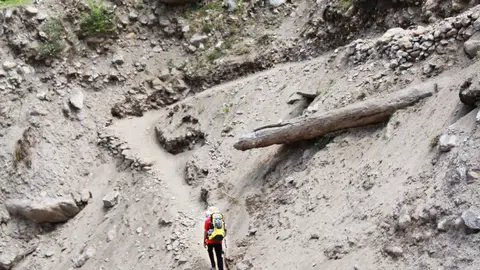Ein einsamer Wanderer mit gelbem Rucksack durchquert einen unebenen Dirt Path inmitten felsigem Bergterrain.