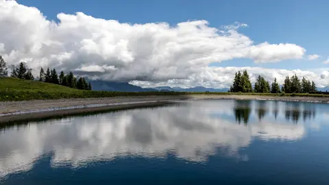 Ein ruhiger See auf dem Land, der dramatische Wolken und Bäume unter einem hellen Himmel spiegelt.