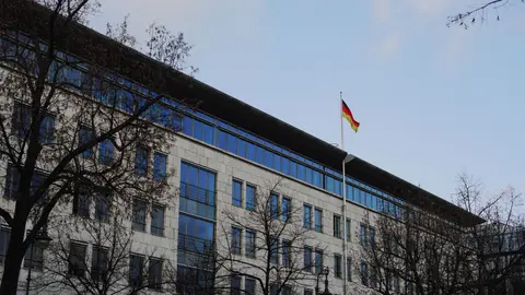 Ein modernes Gebäude mit einer deutschen Flagge, umgeben von kahlen Bäumen unter einem klaren Himmel.